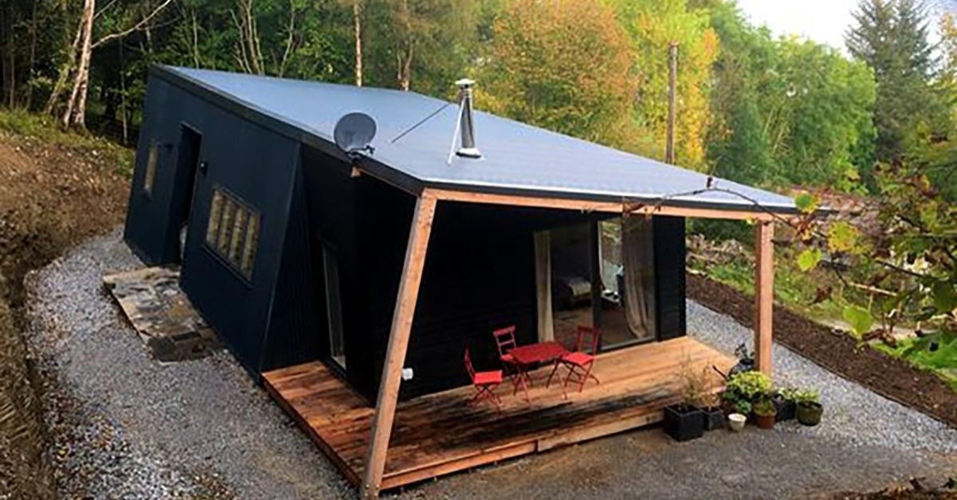 A compact off-grid style cabin home with a wooden deck, surrounded by trees